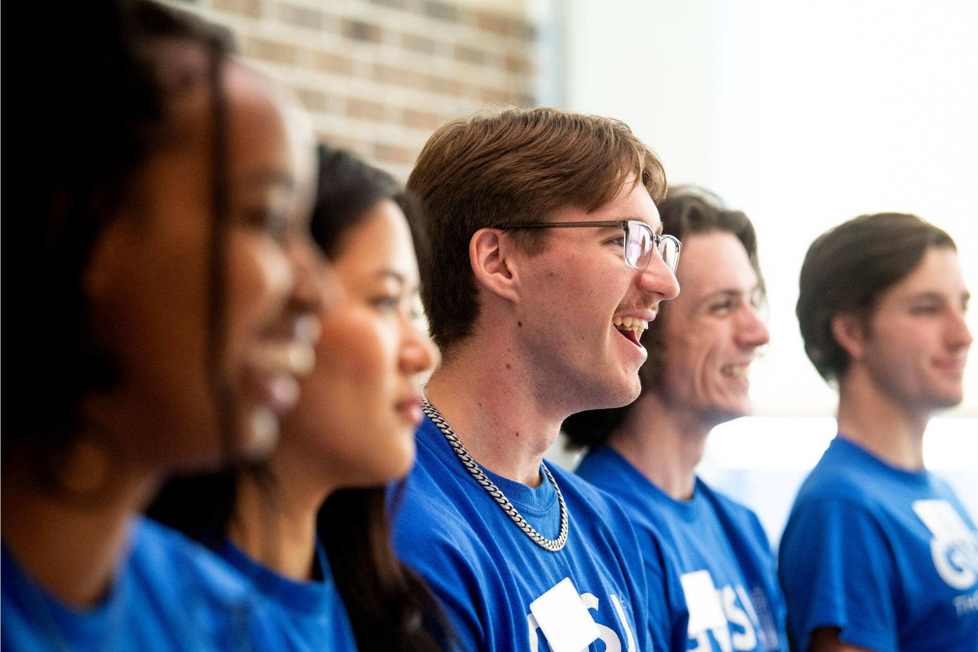 Students wearing blue Thompson Scholar t-shirts smiling with sunlight pouring in from the window behind them.